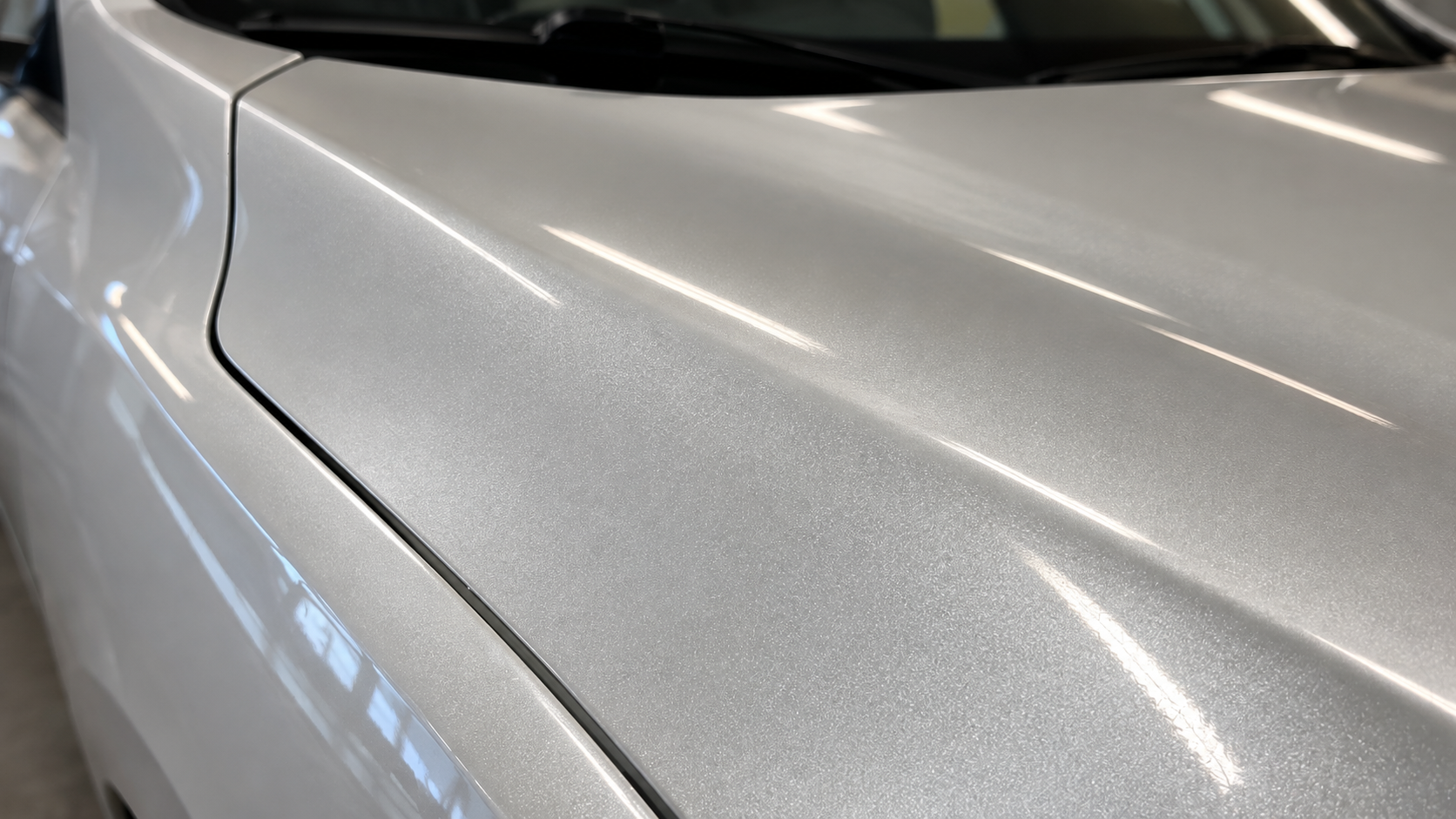 Close-up of a silver metallic car hood and fender showing a smooth paint finish with soft light reflections.